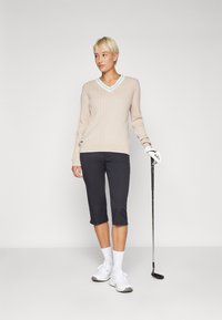 Béžový rebrovaný sveter s svetlomodrým výstrihom do V, kombinovaný s tmavými skrátenými nohavicami a bielymi teniskami, drží golfovú palicu.