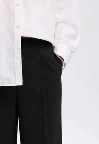 Bijela košulja na kopčanje uparena s crnim krojenim hlačama. Košulja ima ravni kroj i duge rukave s podvrnutim manžetama.