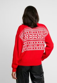Red knitted sweater featuring a white geometric pattern and the phrase "Joyeux Noël" across the back. Long sleeves and ribbed hem.