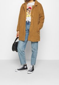 Brązowy, puszysty, oversize'owy płaszcz z polaru z klapowym kołnierzem; noszony na koszulce z nadrukiem, zestawiony z luźnymi niebieskimi dżinsami i czarnymi wysokimi trampkami.