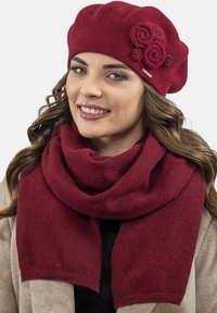 Berretto bordeaux con accenti floreali decorativi abbinato a una sciarpa coordinata, entrambi in tessuto morbido e texturizzato. La sciarpa cade in modo morbido.