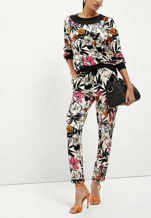 Vrouw draagt bijpassende top en broek met bloemmotief in zwart, wit, roze en oranje, houdt een zwarte handtas vast en draagt oranje sandalen met hoge hakken.