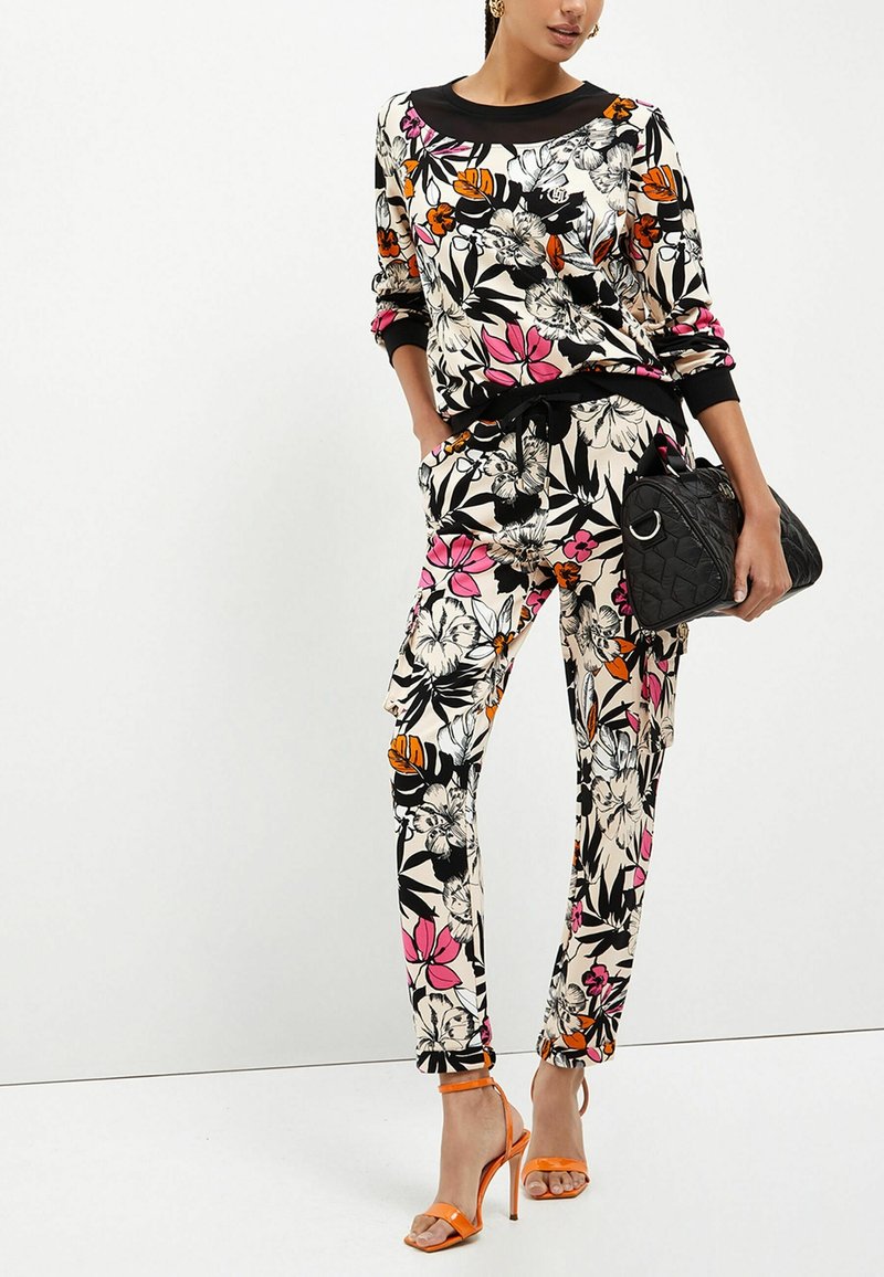 Femme portant un haut et un pantalon à motif floral assorti en noir, blanc, rose et orange, tenant un sac à main noir et portant des sandales à talons hauts orange.