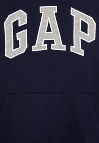 Donkerblauwe sweatshirt met de tekst "GAP" in grote, witte, geborduurde letters. Het heeft een voorzak en is gemaakt van een zachte, gestructureerde stof.