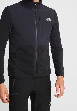 Mand iført sort North Face lynlåsjakke og sorte bukser, stående med armene afslappet mod en lys baggrund.