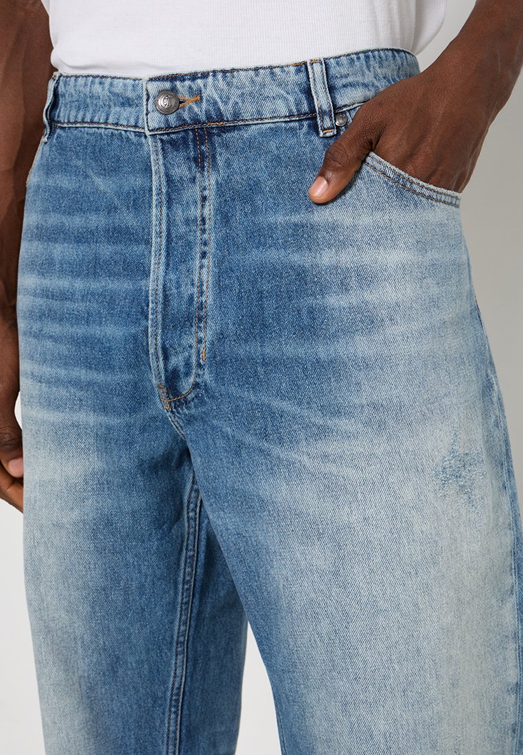 Gros plan sur une personne portant un jean bleu délavé avec une main dans la poche avant et une chemise blanche rentrée.
