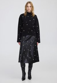 Maglione nero a maglia con decorazioni argentate, abbinato a una gonna in pelle nera. La gonna presenta un design dritto e una texture lucida. Stivali neri al ginocchio.