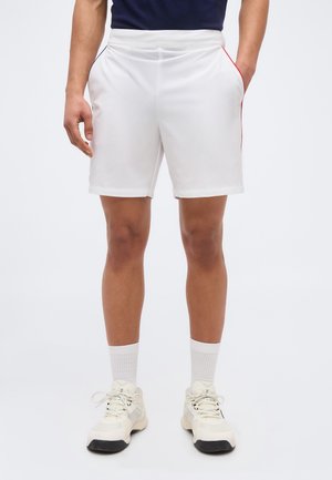 Mężczyzna w białych sportowych szortach z bocznymi kieszeniami, białych skarpetkach i białych tenisówkach stoi na jednolitym tle.