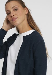 Cardigan en tricot bleu marine avec un motif de points texturé, à ouverture sur le devant et des poignets côtelés, porté par-dessus un t-shirt blanc.