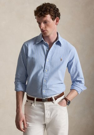 Jeune homme aux cheveux bouclés portant une chemise bleue à rayures boutonnée, un pantalon blanc, une ceinture marron, une montre au poignet et posant avec une main dans la poche.