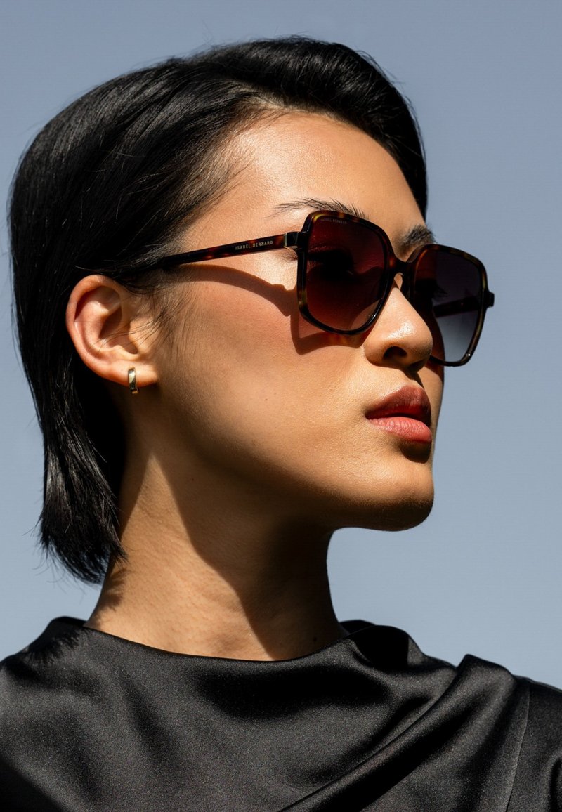  Schildpadzonnebril met donkere lenzen, vierkant frame en dunne pootjes. Het model draagt een zwarte satin top, met kort, donker haar.