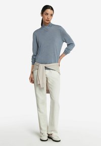 Jasnoniebieski sweter z wysokim kołnierzem, zestawiony z białymi spodniami i beżowym swetrem przewiązanym w talii. Model ma na sobie białe mokasyny.