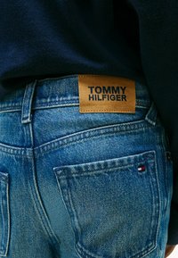 Pantalones cortos de mezclilla en azul deslavado con un parche de cuero marrón etiquetado "TOMMY HILFIGER." Presenta dos bolsillos traseros con un pequeño acento de logo tricolor.