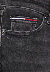 Jeans de mezclilla negra con un bolsillo frontal con detalles a rayas en rojo, blanco y azul, bot�ón de metal y textura de costura visible.