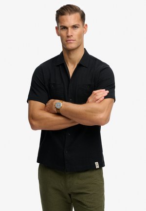 Hombre con cabello corto, vestido con una camisa negra de manga corta abotonada y pantalones verde oliva, con los brazos cruzados y un reloj en la muñeca izquierda.