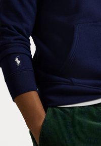 Marinblå sweatshirt med kängurufickor, ribbad mudd och en vit broderad logotyp på ärmen; matchas med gröna manchesterbyxor.