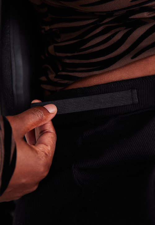 Hand, die den elastischen Bund der schwarzen Hose anpasst, während ein braun-gestreiftes Hemd getragen wird.