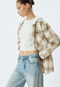 Kariertes Button-up-Hemd in Creme und Braun über einem weißen Cropped-Top, kombiniert mit hellblauen hochgeschnittenen Jeans. Glatte Texturen; lässiges Design.