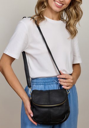 Mustast nahast diivanikott tekstuurse viimistlusega, kuldse luku ja reguleeritava rihmaga, mida hoiab inimene vabaaja riietuses.