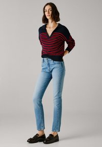 Röstripeeritud tumesinine ja punane kudum, V-kaelusega, koos helesiniste teksadega. Mudel kannab tumedaid mokkasid ja seisab neutraalses taustas.