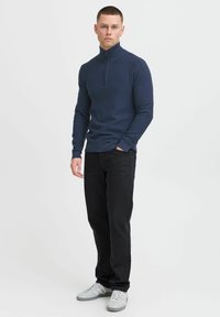 Mornarsko modra pulover s zapenjanjem na vratu in rebrasto strukturo, v kombinaciji s črnimi kavbojkami in sivimi tenisicami. Model stoji pokončno, roke ima v žepih.