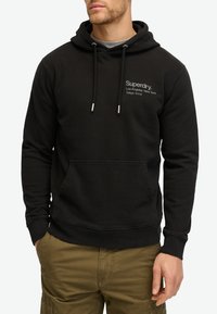 Sudadera con capucha negra de algodón, que cuenta con un bolsillo frontal y detalles en plata en los cordones. El texto en la parte delantera muestra el nombre de la marca y sus ubicaciones.