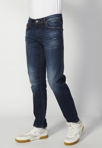 Sötétkék farmernadrág, slim fit szabású, világos kopásokkal, varrott díszítésekkel és klasszikus öt zsebes kialakítással.