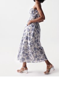 Valkoinen olkaimeton maksimekko, jossa sininen kukkakuvio, kuminauhalla koristeltu yläosa ja sivutaskut, yhdistettynä valkoisiin remmikkäisiin korkokenkiin.