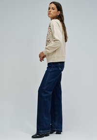 Mujer de pie de lado vistiendo un sudadera beige con bordado floral, jeans de pierna ancha azul oscuro y botas de tacón negras sobre un fondo gris.