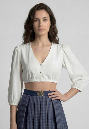 Frau mit welligem braunem Haar, die eine weiße bauchfreie Bluse mit Knöpfen und einen blauen plissierten Rock mit dekorativer Gürtelschnalle trägt.