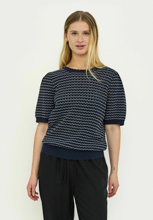 Tengerészkék kötött pulóver hullámmintával, rövid púpos ujjakkal, bordázott nyakkivágással és szegéllyel, sötét, bő szabású nadrággal párosítva.