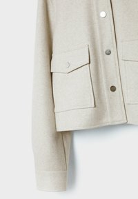 Beige Wolljacke mit langen Ärmeln, zwei Fronttaschen mit Klappe und silbernen Druckknöpfen sowie silberner Druckknopfverschluss vorne.