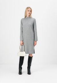 Siva pletena obleka z visokim ovratnikom in dolgimi rokavi, v kombinaciji z črnimi čevlji do kolen in majhno bež torbico.