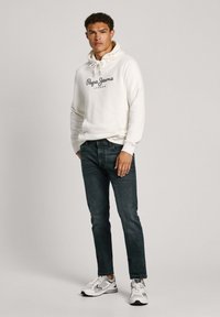 Valge kapuutsiga särkk sildiga "Pepe Jeans London", kombineeritud tumedate siniste teksade ja hallide spordijalatsitega. Sile kangas, vabalt istuv.