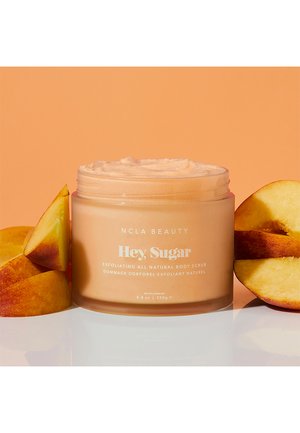 NCLA Beauty Hey Sugar body scrub in a clear jar with a light peach color, surrounded by peach slices, highlighting its natural exfoliating texture.