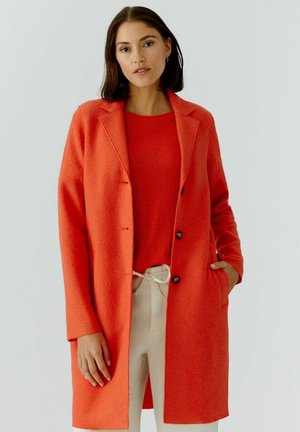 Frau mit dunklen Haaren, die einen leuchtend orangefarbenen Mantel und Pullover sowie beige Hosen mit Kordelzug trägt, steht vor einem einfarbigen hellen Hintergrund.