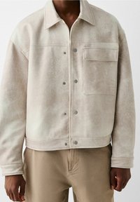 Beige keinonahkainen takki, jossa edessä napitus, rintatasku ja lyhyt malli. Siinä on klassinen kaulus ja painonappikoristeet.