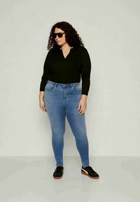 Μαύρη μακρυμάνικη μπλούζα συνδυασμένη με ανοιχτό μπλε στενές τζιν. Το μοντέλο φοράει γυαλιά ηλίου και μαύρα αθλητικά παπούτσια με καφέ σόλες. Ορατά σγουρά μαλλιά.