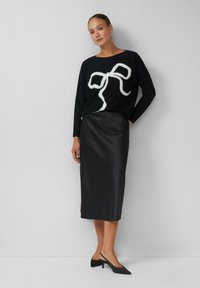 Kobieta w czarnej bluzie z białym abstrakcyjnym wzorem w kształcie kokardy, czarnej spódnicy midi i czarnych butach na obcasie typu slingback, stojąca przed jasnoszarym murem.