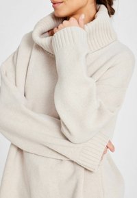 Lichtbeige coltrui met geribbelde details, brede manchetten, een ontspannen pasvorm en een zachte textuur, gedeeltelijk weergegeven met handen.