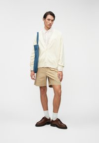 Κρέμα πουλόβερ με πλέξη καλωδίων, ανοιχτόχρωμο πουκάμισο με ρίγες, μπεζ σορτς, καφέ δερμάτινα παπούτσια, σε συνδυασμό με μπλε τζιν τσάντα.