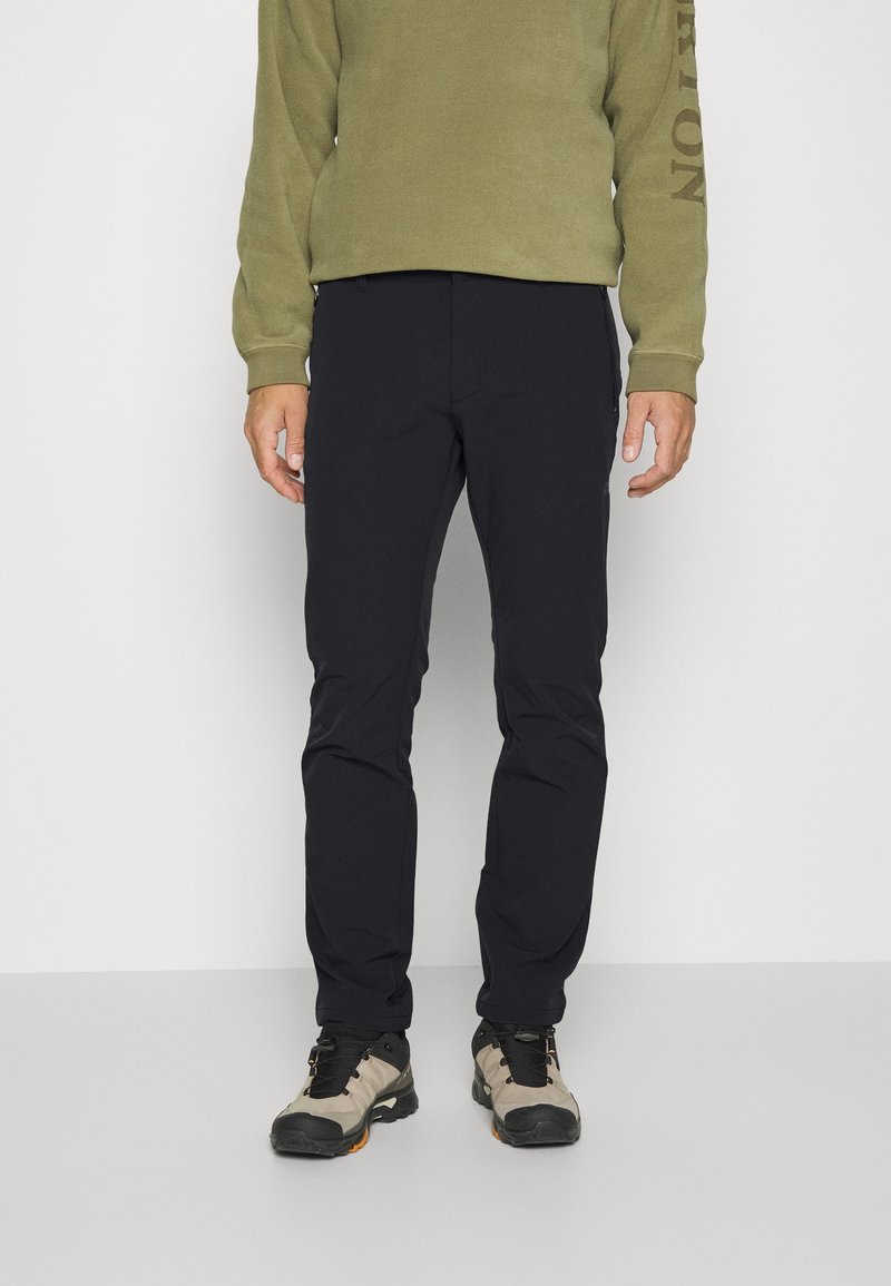 Pantalons extensibles gris foncé avec une coupe slim, associés à un sweat-shirt à manches longues vert foncé avec du texte sur la manche, et des chaussures de randonnée beiges.