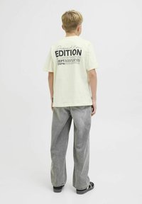 Camiseta de algodón amarilla clara con un estampado gráfico en la parte trasera que presenta texto en negrita negro. Combinada con jeans de pierna ancha grises y zapatos negros.