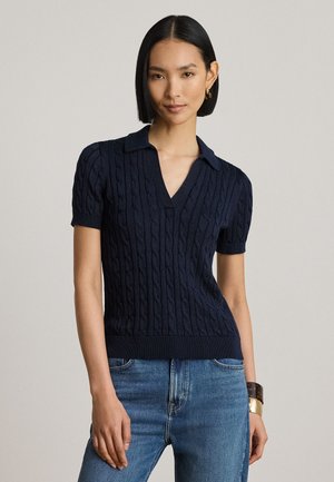 Kvinde med glat sort hår iført en marineblå kortærmet kabelstrik sweater og blå jeans, stående mod en ensfarvet baggrund.
