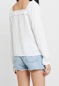Blouse blanche à manches longues avec un décolleté carré, dotée de dentelle. Associée à un short en denim bleu clair avec des bords effilochés.