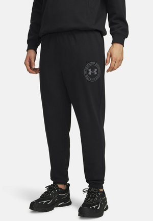Mann trägt schwarze Jogginghose mit Under Armour-Logo am linken Oberschenkel und schwarze Sportschuhe vor einfachem Hintergrund.