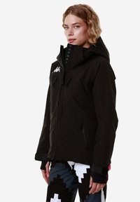 Giacca da sci nera con colletto alto, tasche con zip e logo sul petto. Presenta un cappuccio e un tessuto texturizzato. Abbinata a pantaloni con fantasia.