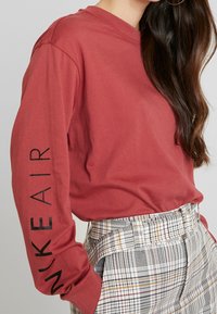 Röd långärmad tröja med "NIKE AIR" tryckt i svart på vänster ärm, kombinerad med rutiga högmidjade byxor med bälte.