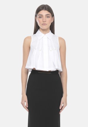 Donna con capelli castani lisci che indossa una camicia bianca plissettata senza maniche e corta e una gonna nera a vita alta, in piedi davanti a uno sfondo semplice.