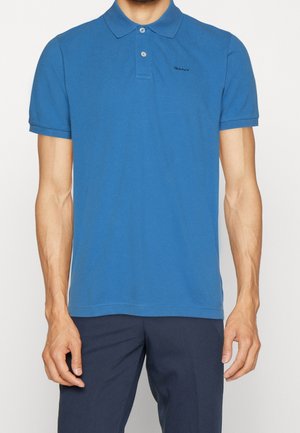 Homme portant un polo bleu à manches courtes avec un col et deux boutons, associé à un pantalon bleu marine foncé sur un fond uni.